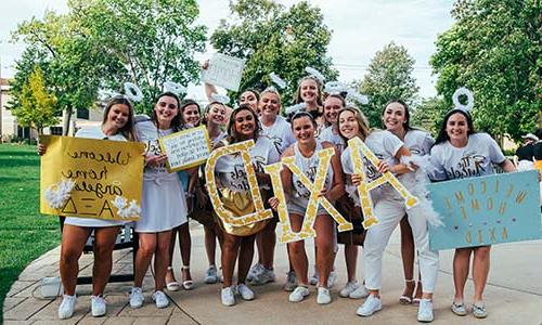 A group of smiling young women dressed in white with angel-themed accessories, 拿着写着“轴”的牌子和字母,可能是欢迎新成员加入她们的姐妹会.